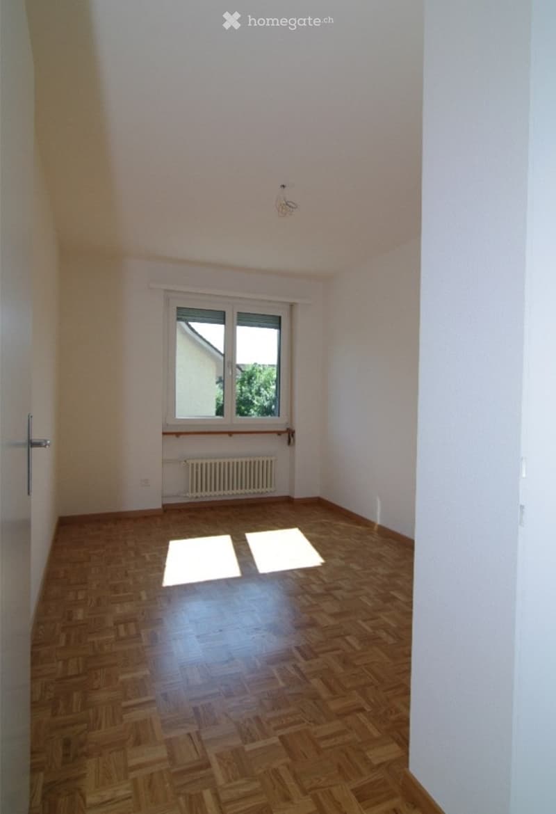 Empty room with wooden parquet flooring, white walls, and a window with a radiator below. No furniture or equipment present.