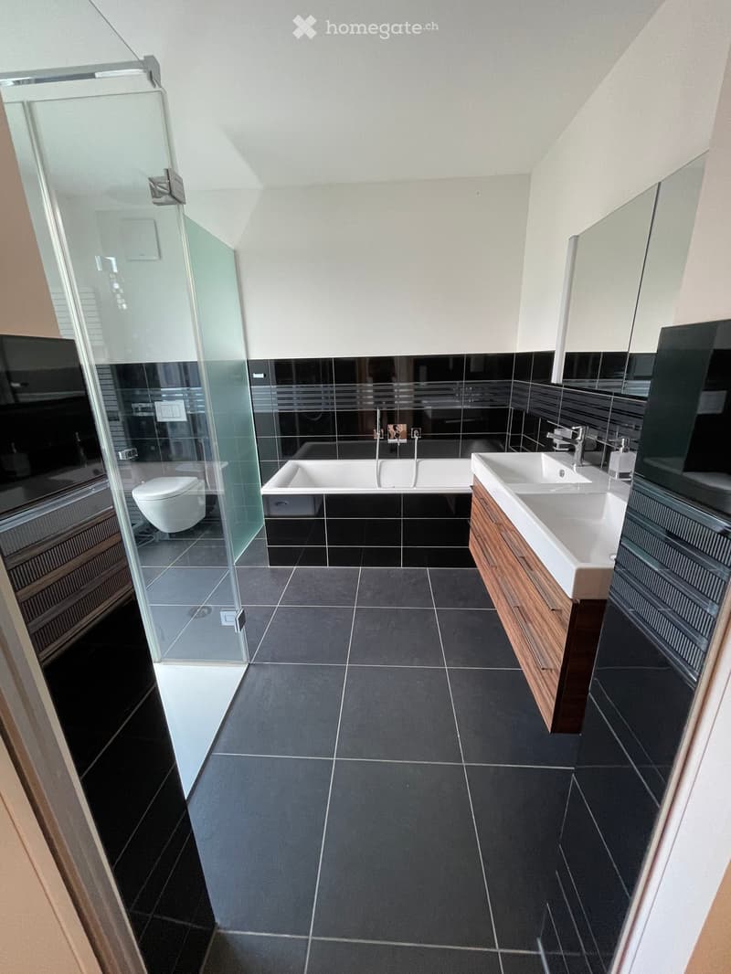 Modern bathroom with dark tiled floor and walls, featuring a bathtub, double sink, and toilet in a glass-enclosed shower area.