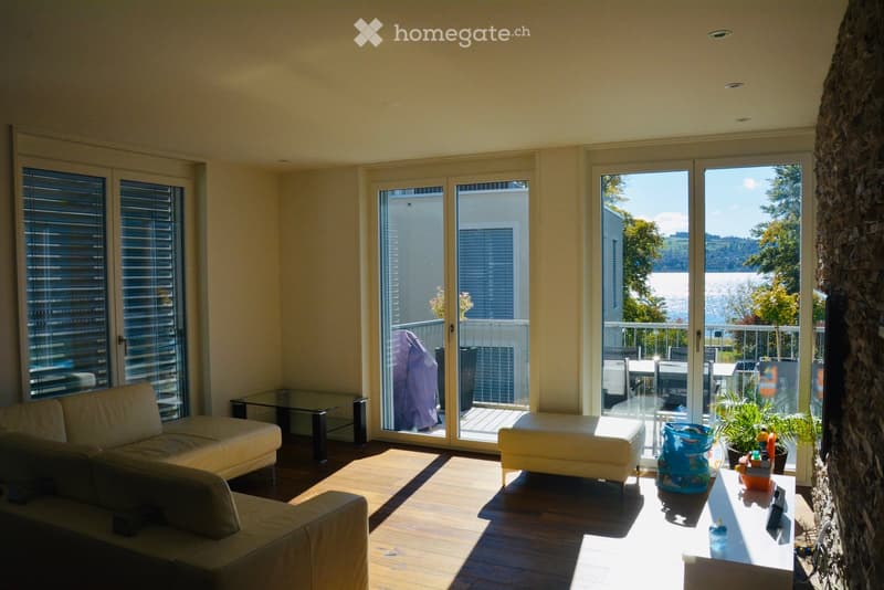 Bright living room with large windows and sliding doors leading to a balcony, featuring a white sofa, ottoman, coffee table, and a small table with a plant.