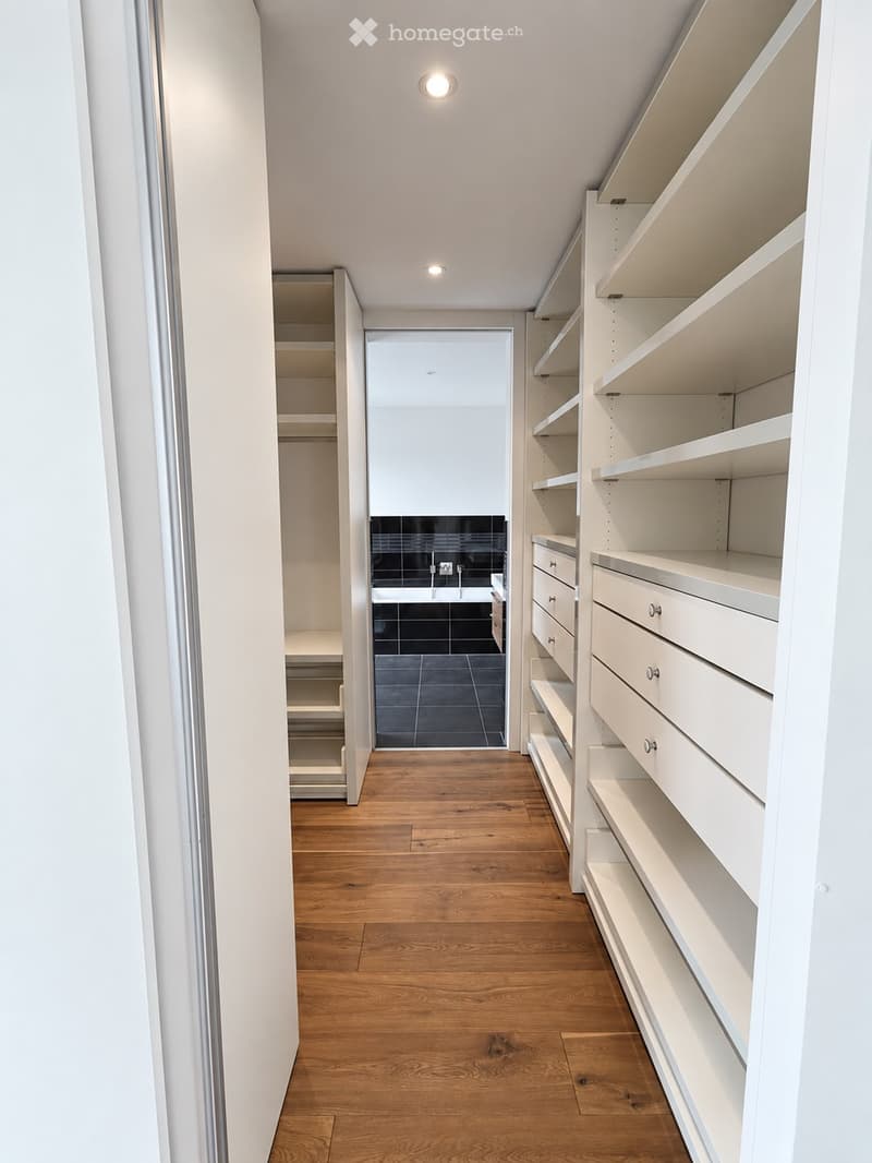 A clean, well-lit hallway with wooden flooring, white built-in shelving units on one side, and an open doorway leading to a bathroom with black tiles and a sink.