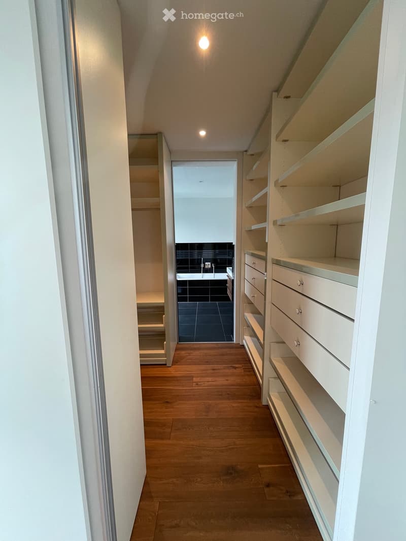 Empty hallway with wooden flooring, white walls, built-in shelving unit on the right, and a view into a bathroom with black tiles and a bathtub.