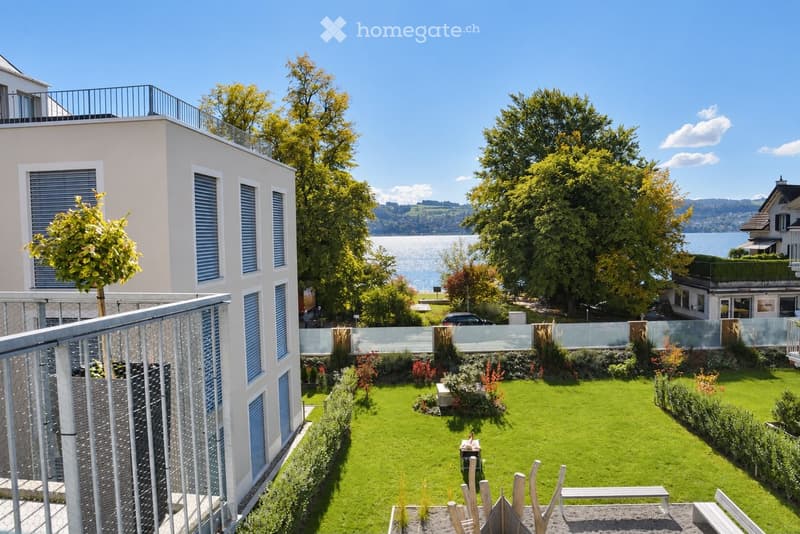 Well-maintained garden with green lawn, small trees, shrubs, and a view of a lake and surrounding landscape.