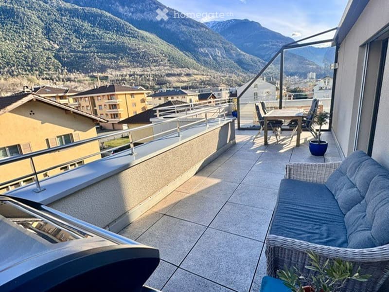 Balcony with tiled floor, metal railing, outdoor furniture including a sofa, table, and chairs, overlooking a scenic mountain view.
