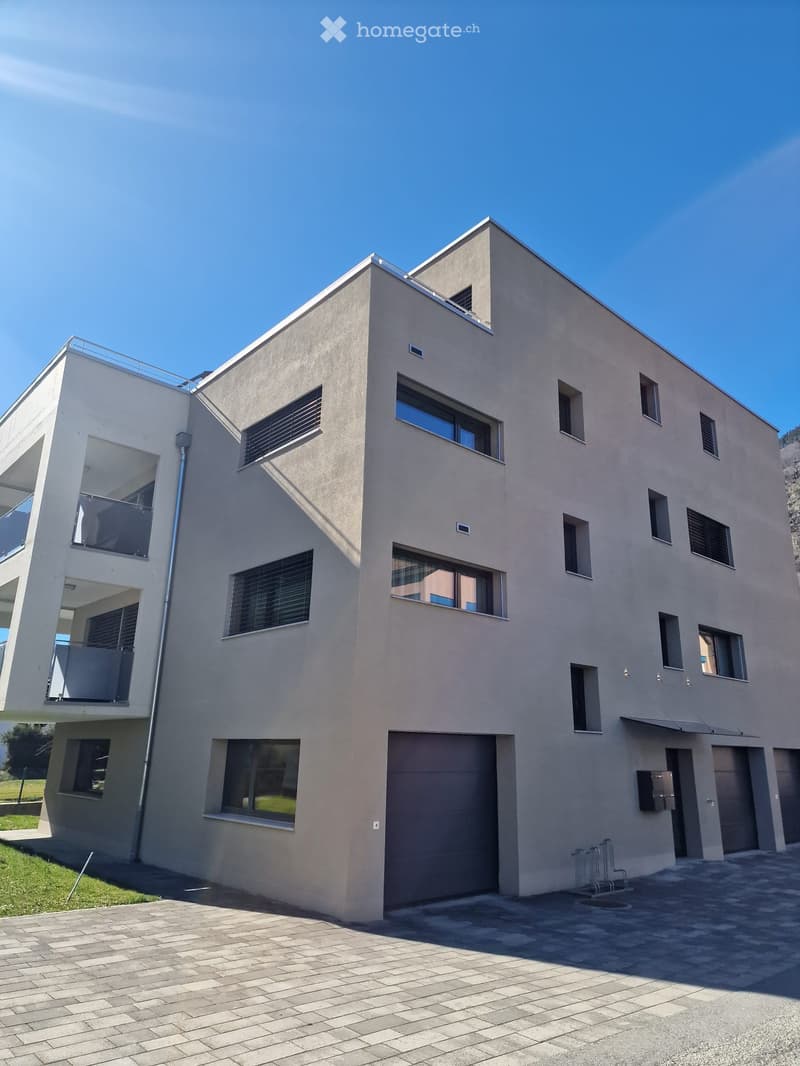 Modern two-story house with beige facade, large windows, and a garage entrance. Features a balcony on the upper level and a paved driveway.