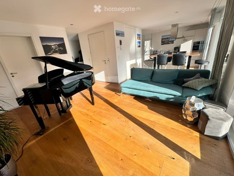 Bright, open-plan living and dining area with wooden flooring, a blue sofa, a black piano, a small round table with chairs, and a modern kitchen area with cabinets and bar stools.