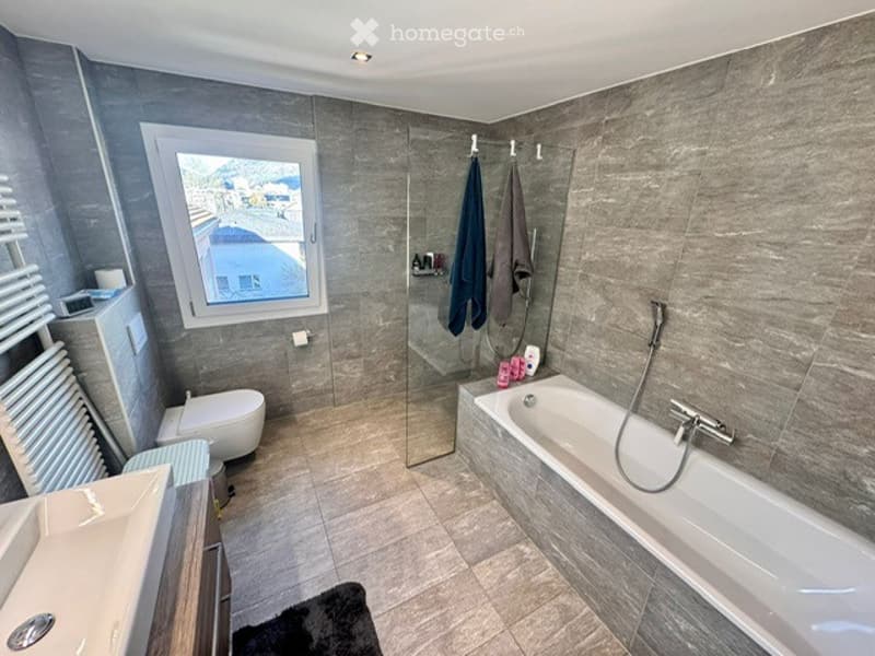 Modern bathroom with grey tiled walls and floor, large window with view, bathtub, toilet, sink, glass shower enclosure, and towel rack with towels.