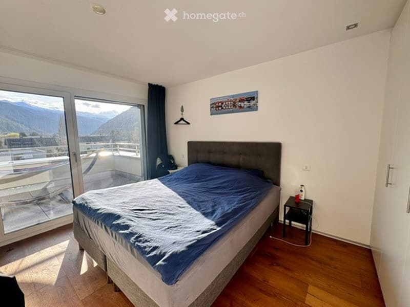 Bright bedroom with wooden flooring, large window offering a mountain view, a bed with blue bedding, a nightstand, and a picture frame on the wall.