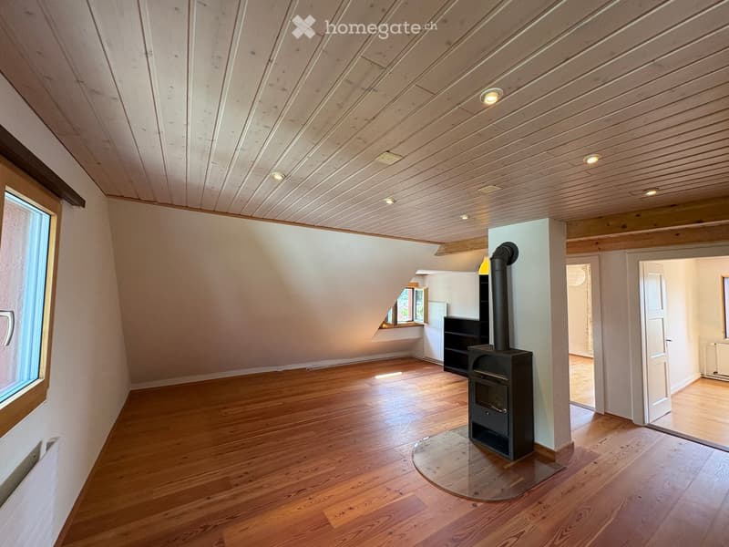An empty attic room with wooden floors, white walls, and a slanted wooden ceiling. Features include a fireplace with a chimney, built-in shelving, and large windows allowing natural light.