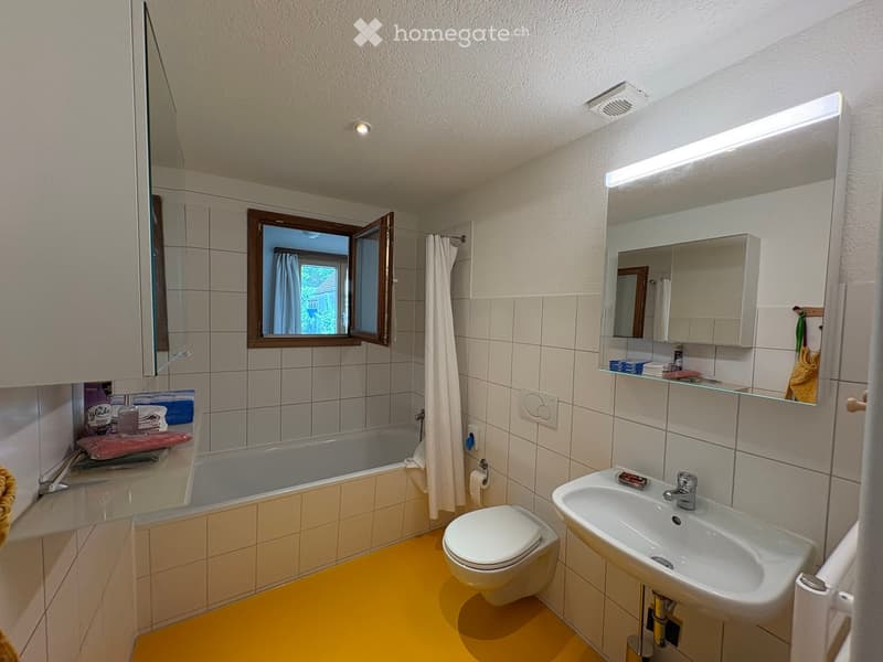 Bathroom with a yellow floor, white tiled walls, a bathtub, toilet, sink, mirror, and a window with a curtain.