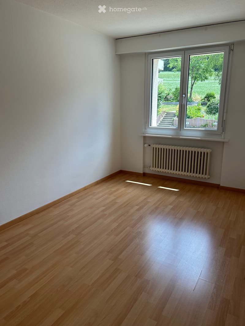 Empty room with light wooden flooring, white walls, a radiator, and a large window offering a view of a garden area.