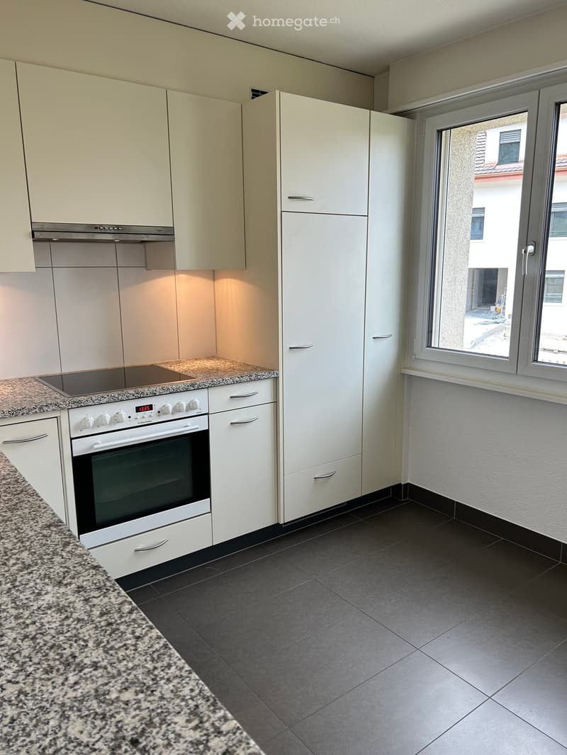 Modern kitchen with white cabinets, granite countertop, built-in oven, and refrigerator. Features large windows with a view of the street.