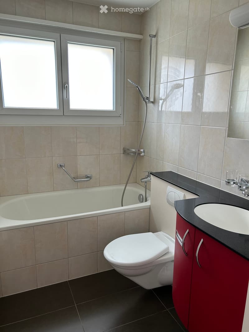 Modern bathroom with beige tiled walls, black countertop, red cabinet, bathtub, toilet, and sink.