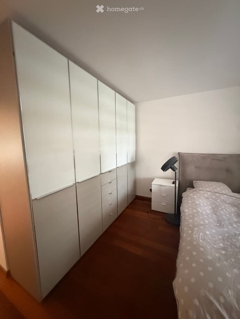 Modern bedroom with a wooden floor, a large mirrored wardrobe, a bed with a tufted headboard, a nightstand, and a floor lamp.