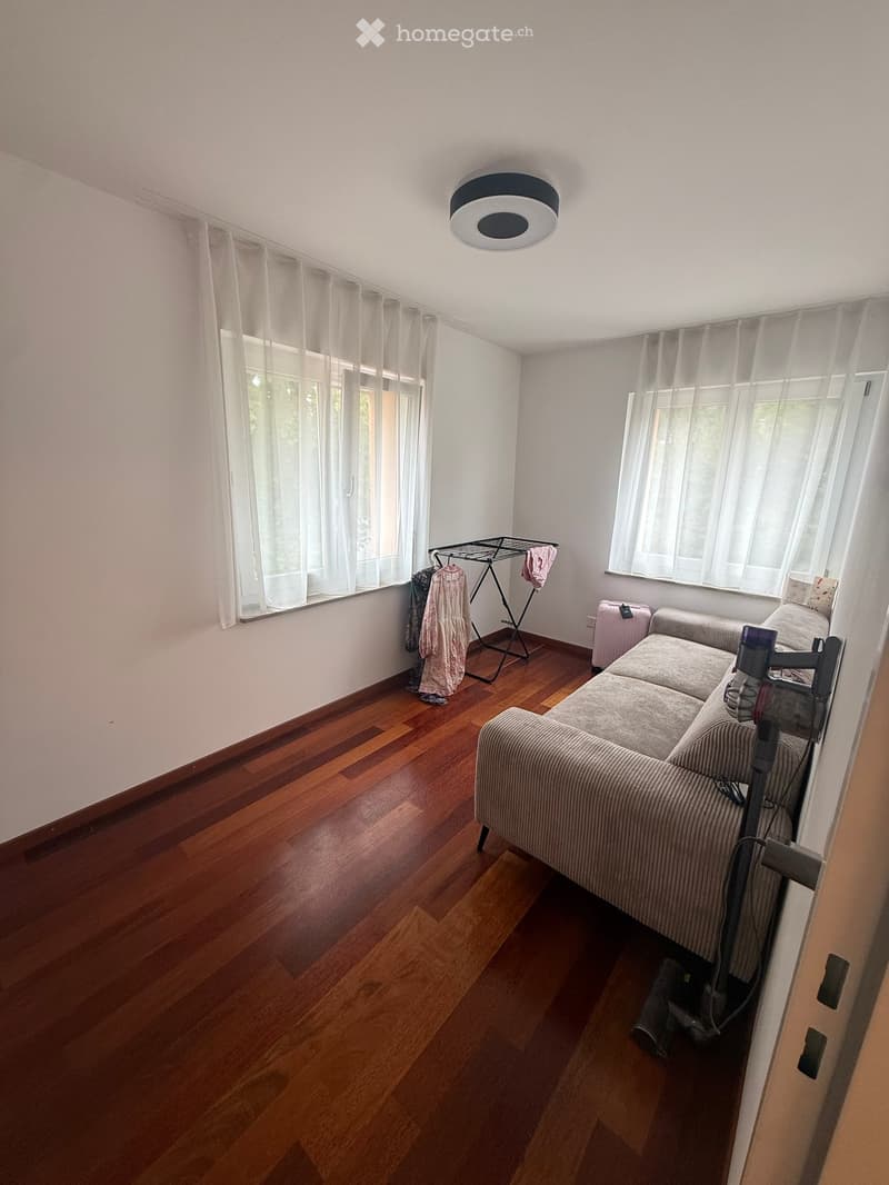 A furnished living room with a grey sofa, ottoman, wooden floor, large windows with white curtains, and a clothing rack with clothes.