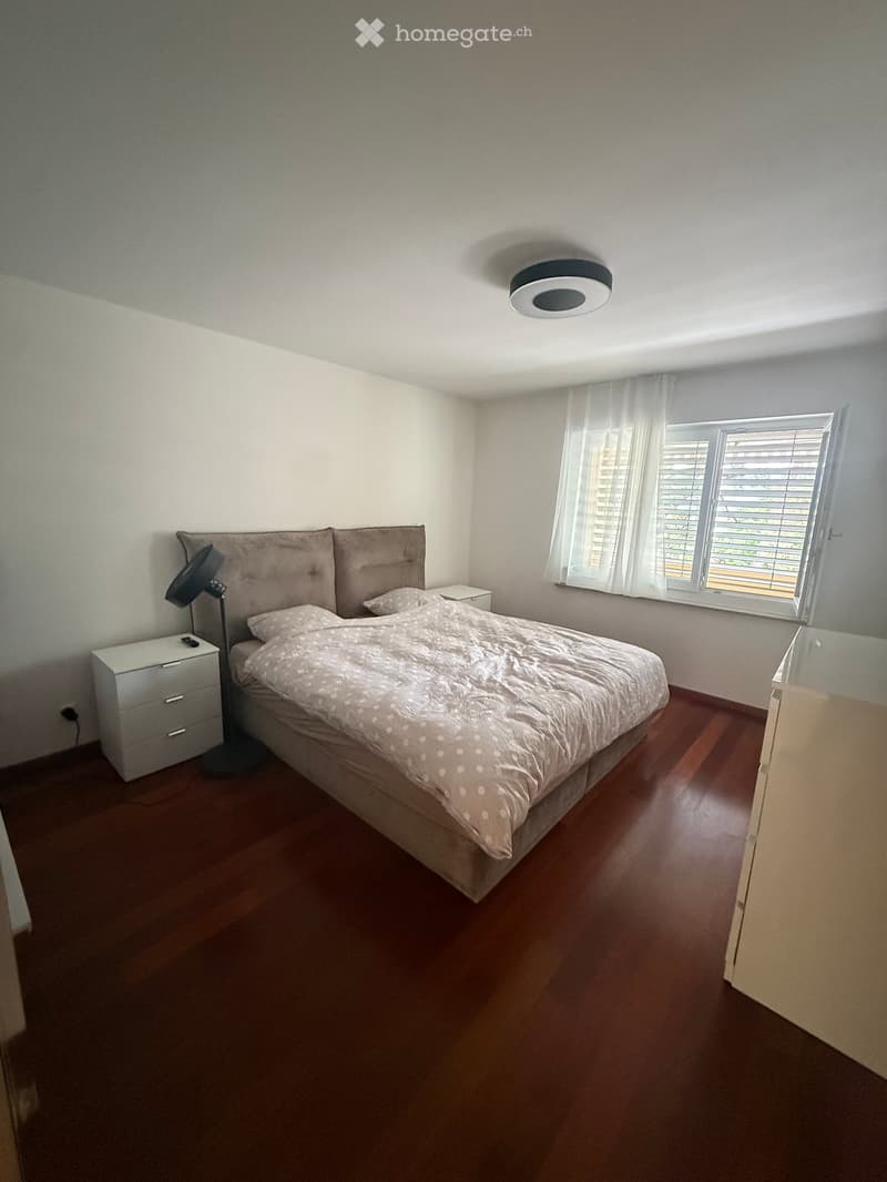 A well-lit bedroom with a wooden floor, a large bed with a patterned duvet, a nightstand, a lamp, and a drawer unit.