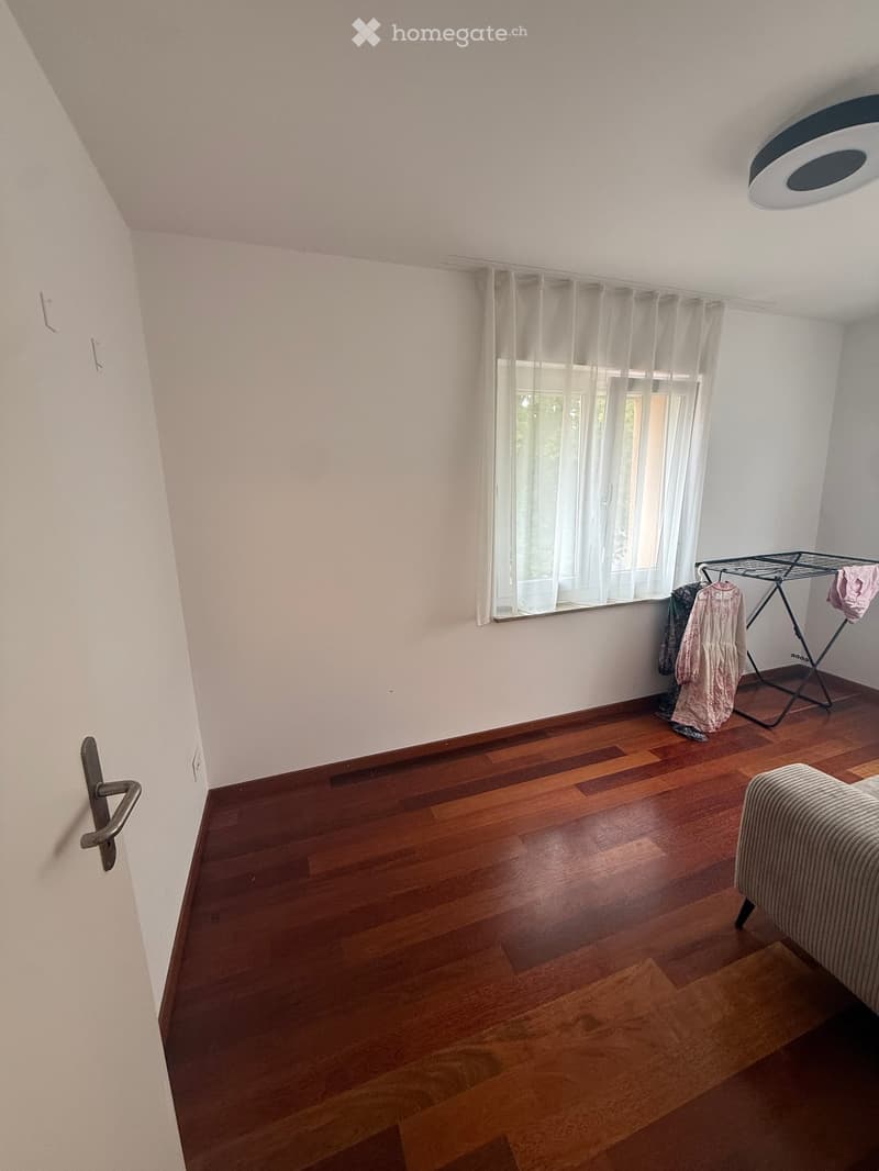 A small, unfurnished room with wooden flooring, white walls, and a large window with white curtains. A sofa is partially visible on the right side, and a clothing rack with a few garments is near the window.