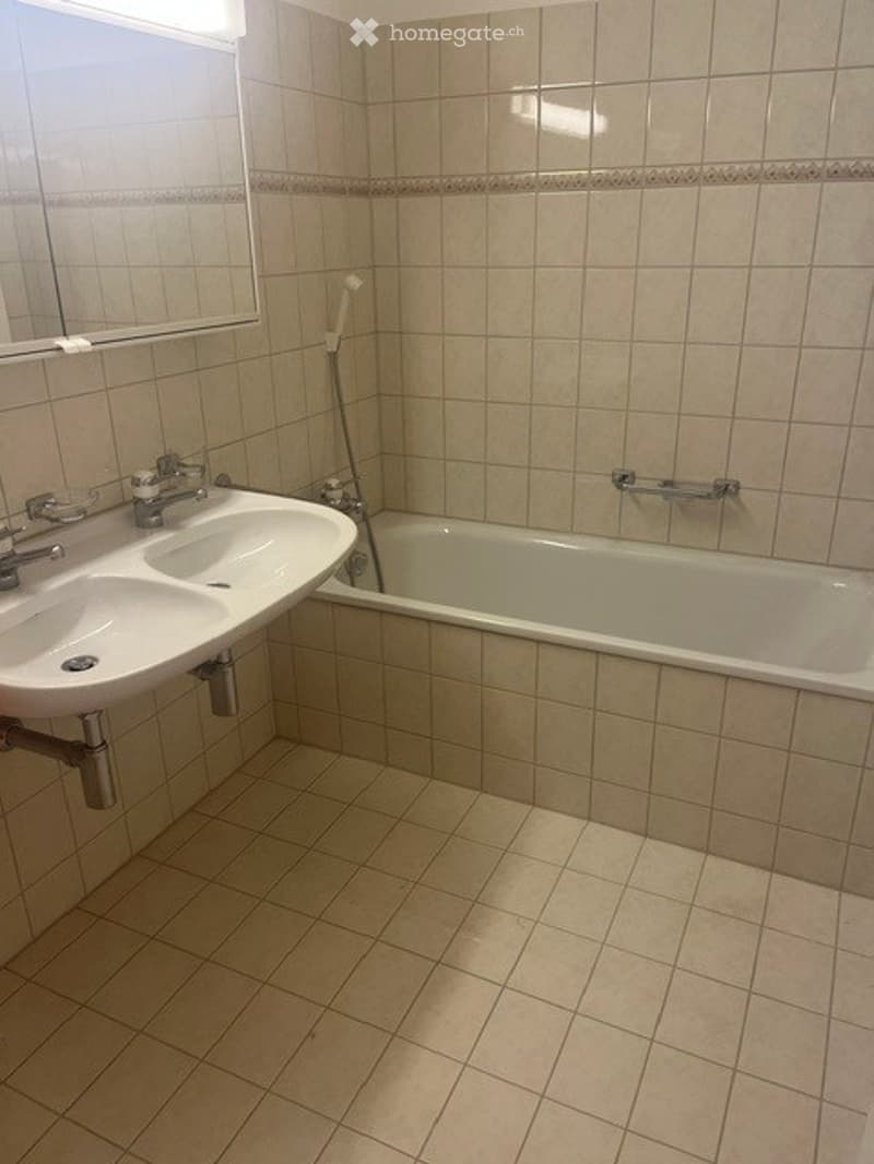 A clean, tiled bathroom with a double sink, bathtub, and a mirror above the sink. The walls and floor are covered in light beige tiles, and the space appears well-maintained.