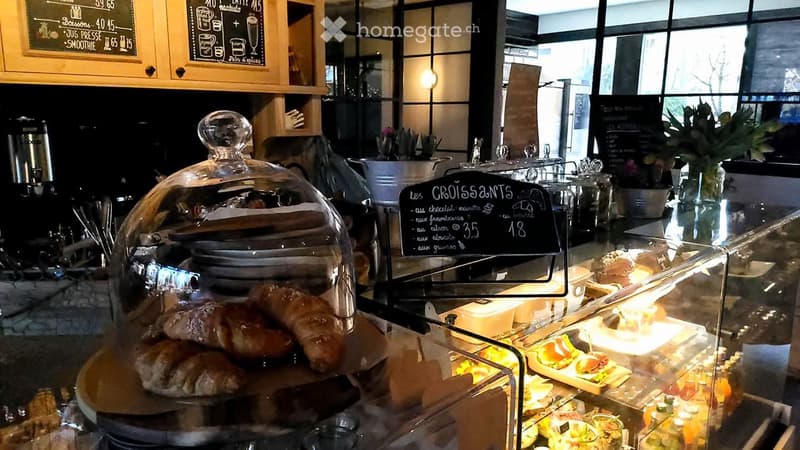 A bakery display case with pastries, a glass dome over croissants, and a menu board listing croissant prices. The counter features a variety of baked goods, including croissants, muffins, and other pastries, with a coffee machine and flower pots in the ba