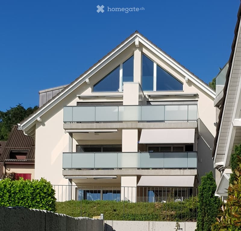 Modern two-story house with white exterior, large windows, and balconies. Features a triangular roof and contemporary design.