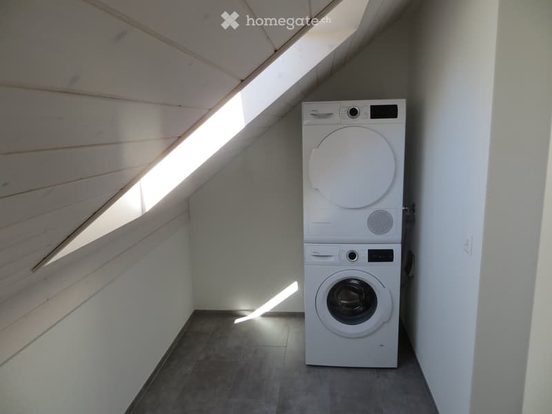 A small, well-lit room with a slanted wooden ceiling and a skylight, containing a stacked washing machine and dryer.