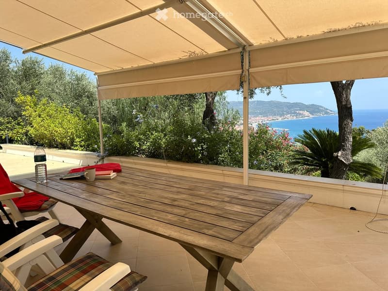 A wooden table and chairs are set up under a beige awning, overlooking a scenic coastal view with trees and the sea.