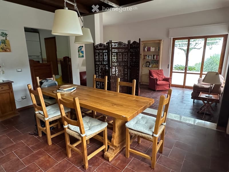 Dining area with a wooden table and chairs, a red armchair, a sofa, a bookshelf, and a decorative screen. The room has tiled flooring and large windows leading to an outdoor area.