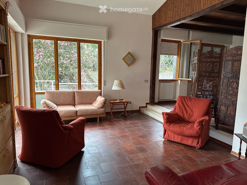 Living room with a beige sofa, red armchairs, a small wooden side table with a lamp, and a decorative screen. The room features wooden flooring and large windows with decorative glass.