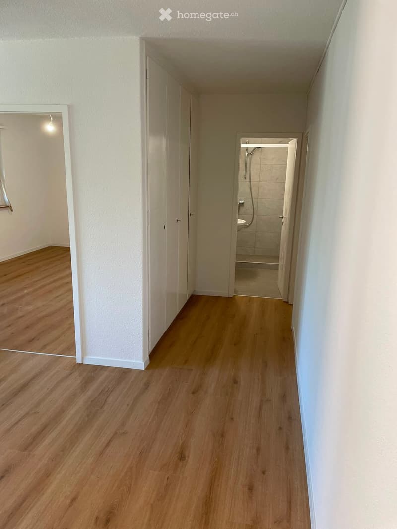 Empty hallway with wooden flooring, white walls, and open doors leading to other rooms, including a bathroom with a shower.