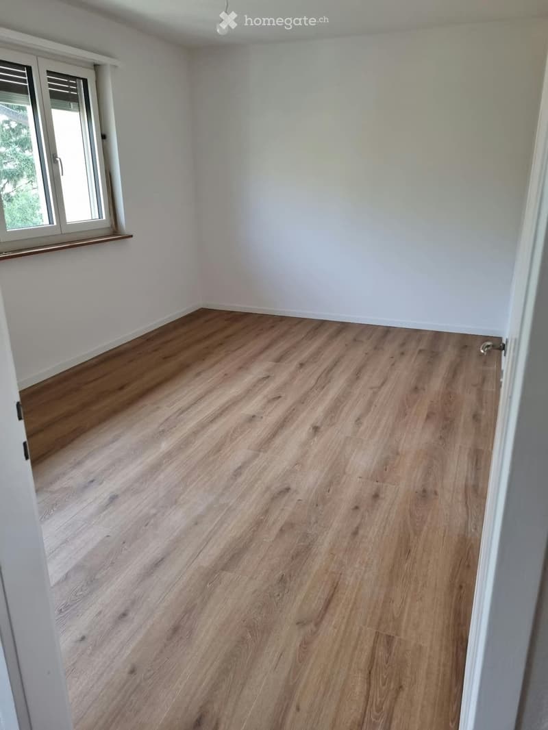 Empty room with light wooden flooring and white walls, featuring a large window with a wooden sill and a sliding glass door leading outside.