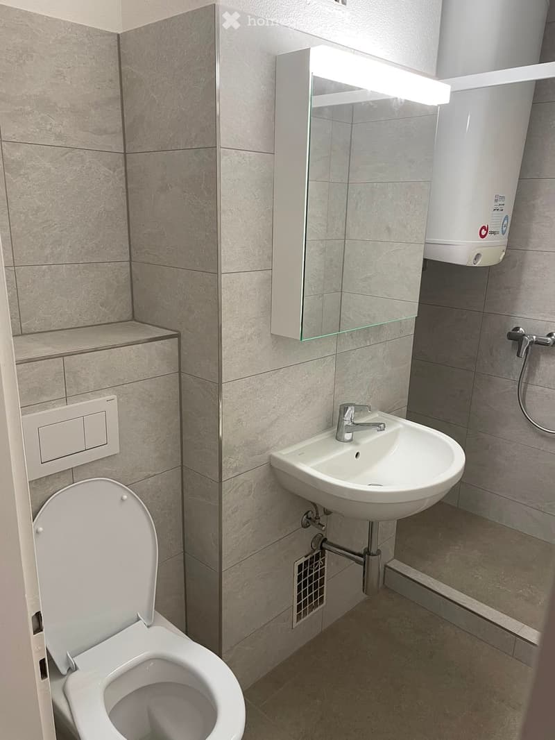 Modern bathroom with a toilet, sink, and mirror. The walls are tiled in a light beige color, and the floor is also tiled. A small shelf is mounted on the wall next to the toilet.