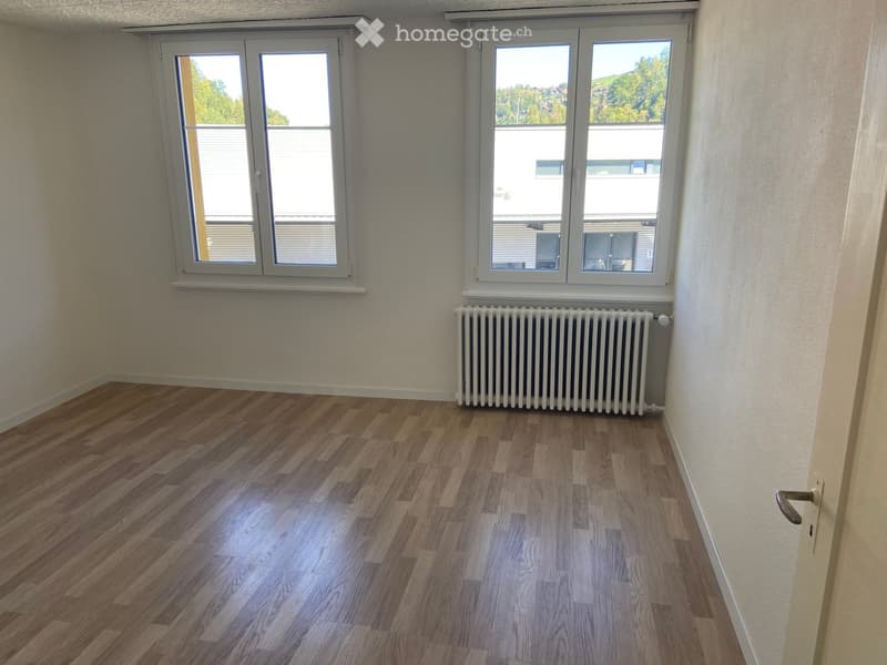 Empty room with wooden flooring, white walls, and large windows with blinds. A radiator is visible under one window.