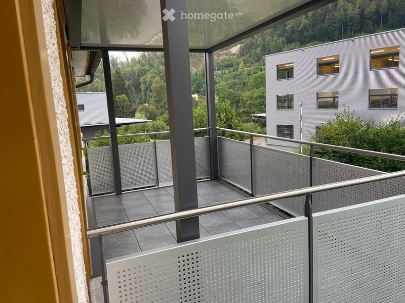 Modern balcony with metal railings, tiled floor, and a view of a forested area and contemporary buildings.