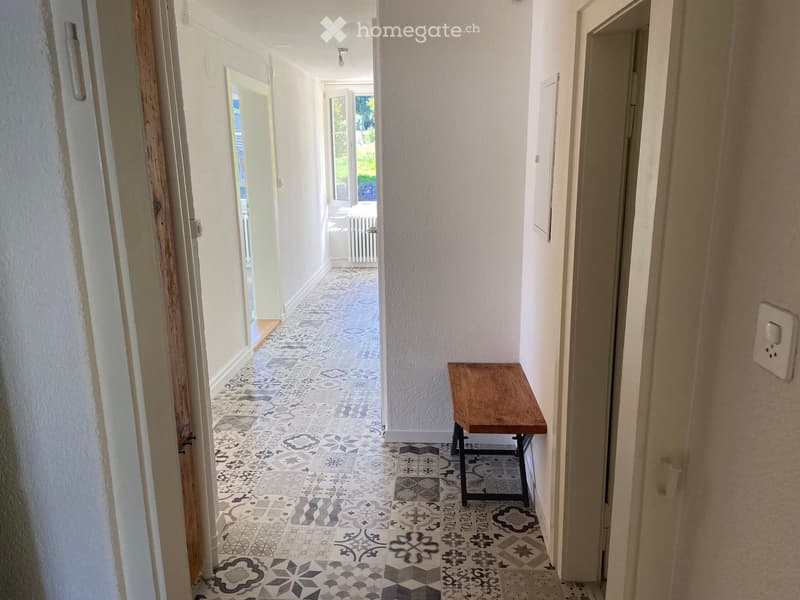 Hallway with patterned tile flooring, white walls, and a small wooden bench placed against the wall.