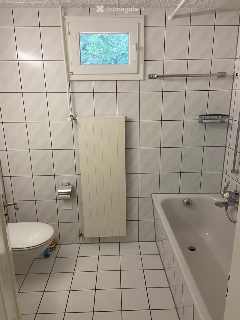 Bathroom with tiled walls and floor, featuring a toilet, bathtub, radiator, and a window above the bathtub.