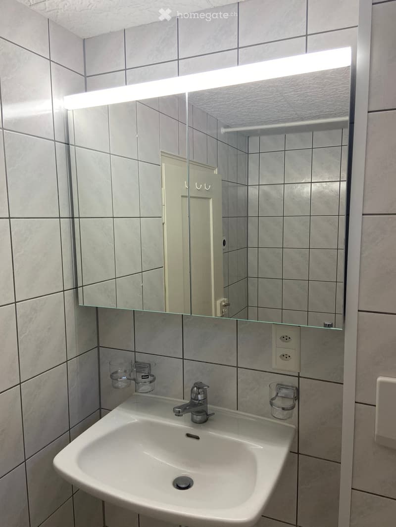 Modern bathroom with a white sink, chrome faucet, and a large mirror with built-in lighting. Three clear cups are placed on the sink counter.