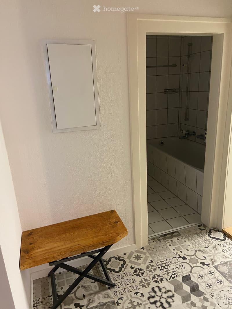 Hallway with patterned tiles, a wooden bench, and an open doorway leading to a tiled bathroom with a bathtub and shower area.