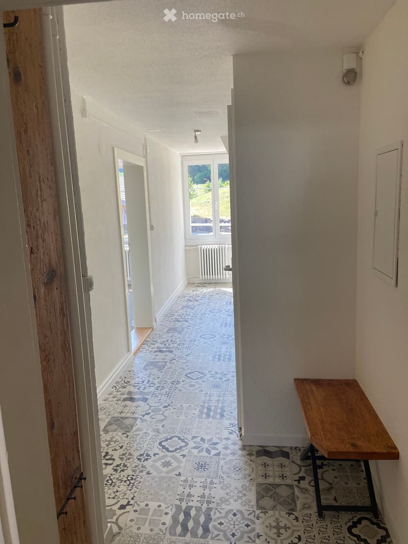 Hallway with patterned tile floor, wooden bench on right side, doors and windows visible.