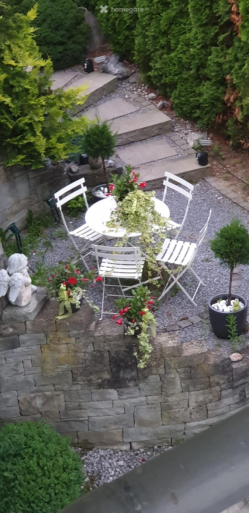 A small, well-maintained garden area with a stone wall, a round white table surrounded by four white chairs, potted plants, and a decorative statue.
