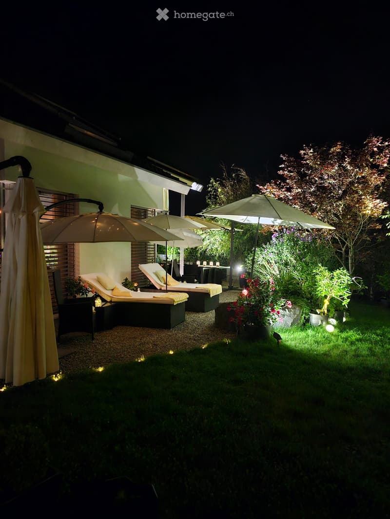 Well-lit outdoor garden area with lounge chairs, umbrellas, and a small table, surrounded by greenery and flowers.