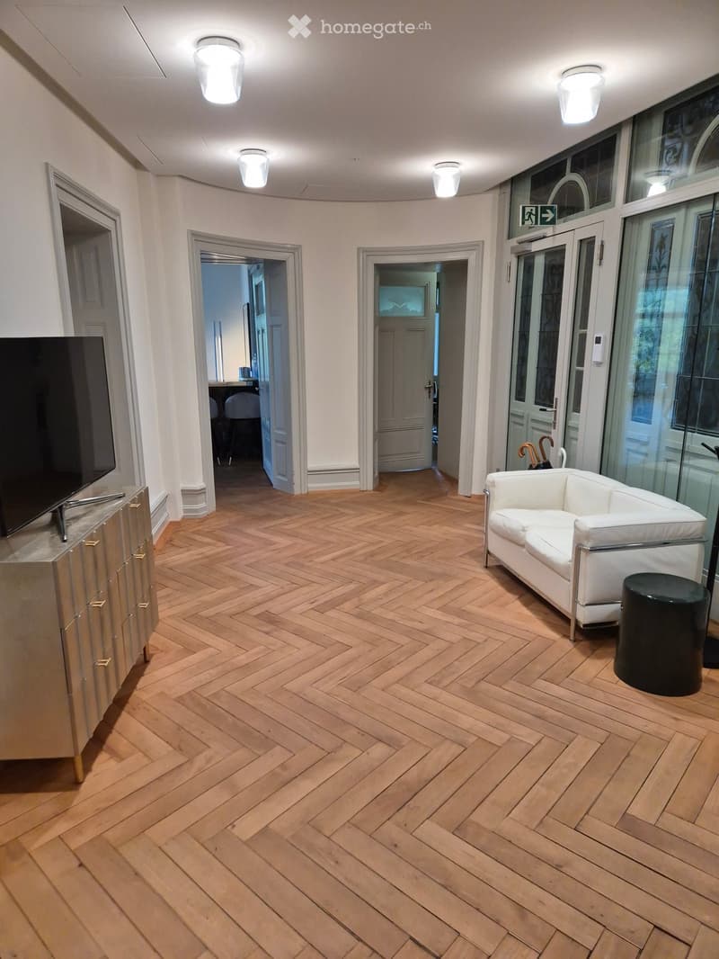 Modern living room with herringbone wooden flooring, white sofa, black stool, and a cabinet with a screen on top.
