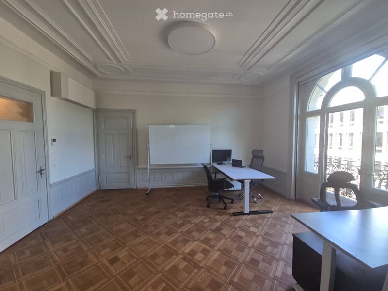 Office space with wooden parquet flooring, large windows, whiteboard, and modern furniture including a table, chairs, and a laptop.