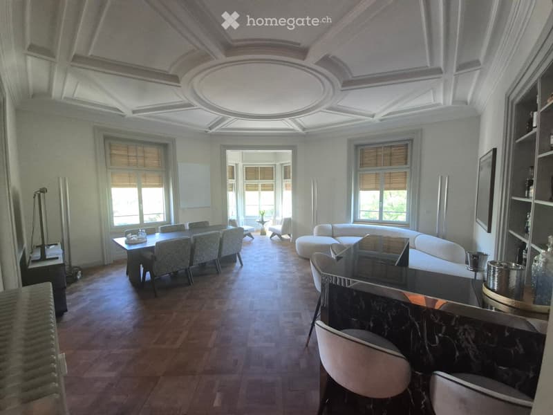 Elegant living and dining area with a high, ornate ceiling, large windows, a white sectional sofa, a dining table with chairs, and a modern bar area with bar chairs and a shelf.