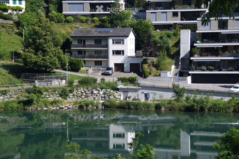 A cluster of modern houses situated on a hillside overlooking a calm river, featuring balconies, large windows, and a mix of contemporary and traditional architectural styles.