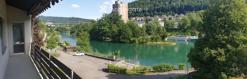 Balcony overlooking a river with a town and hills in the background, featuring a parking area and a small shed.