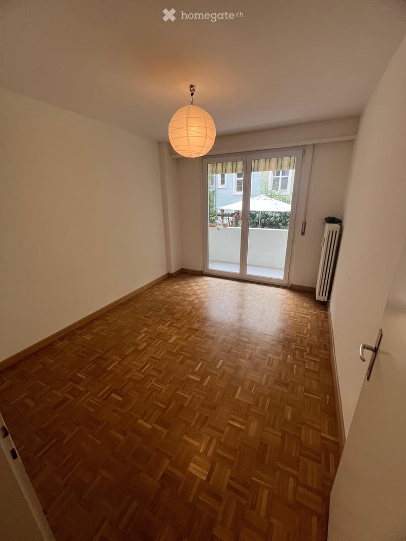 Empty room with wooden herringbone flooring, white walls, and a large sliding glass door leading to a balcony. A single hanging light fixture is visible.