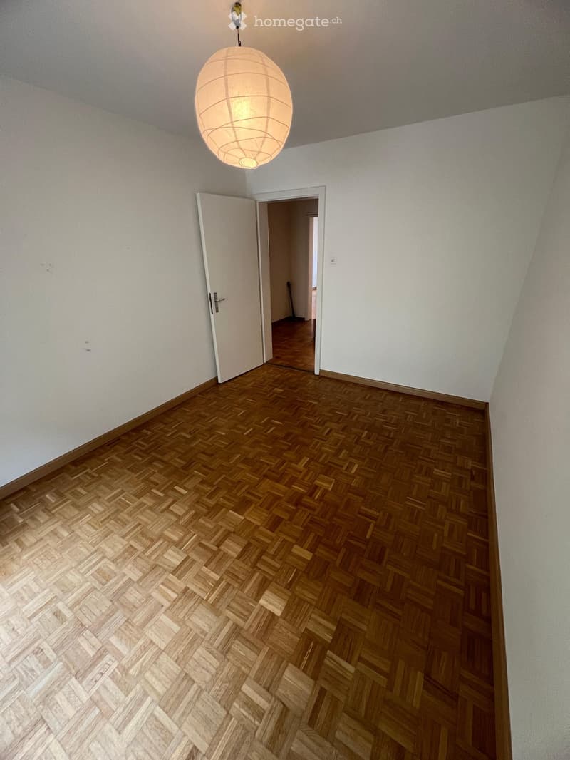 Empty room with wooden herringbone flooring, white walls, and a single hanging light fixture. An open doorway leads to another room.