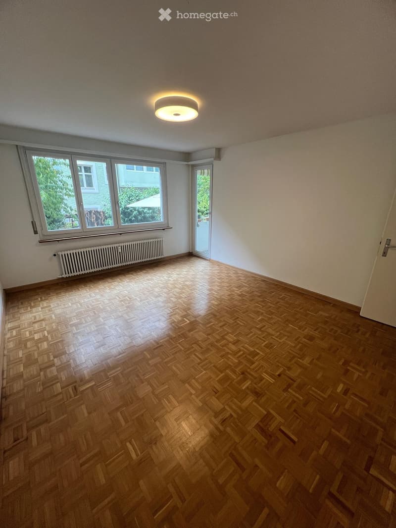 Empty room with wooden herringbone flooring, white walls, large windows, and a ceiling light fixture.