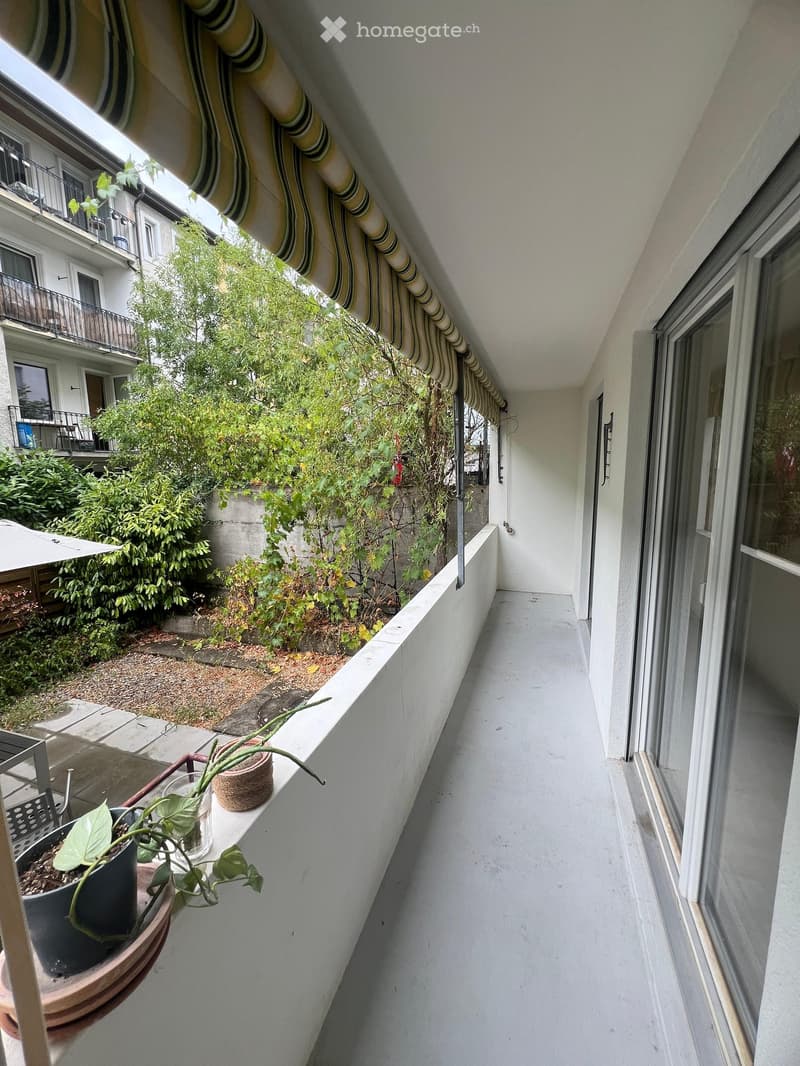 Balcony with white floor, potted plants, striped awning, and view of neighboring building and greenery.