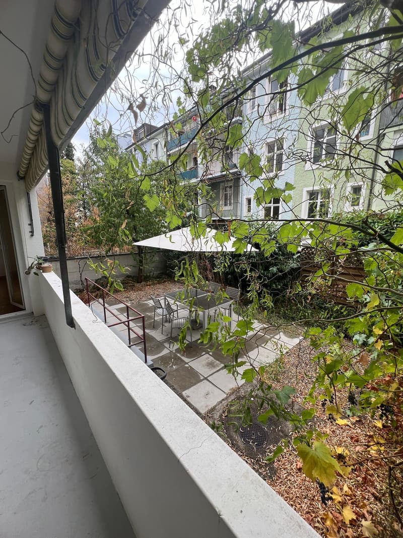 Balcony overlooking a garden area with outdoor furniture, surrounded by climbing plants and neighboring buildings.