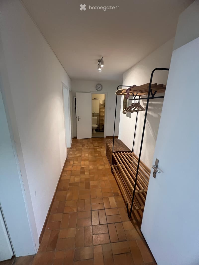 A narrow hallway with wooden tiled flooring, white walls, and a slanted ceiling. A coat rack with hangers is visible on the right side, and a clock is mounted on the wall. An open door leads to a bathroom with a toilet and sink visible.